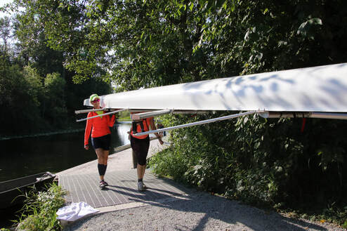 Two rowers are shown helping carry a boat from it's bow.