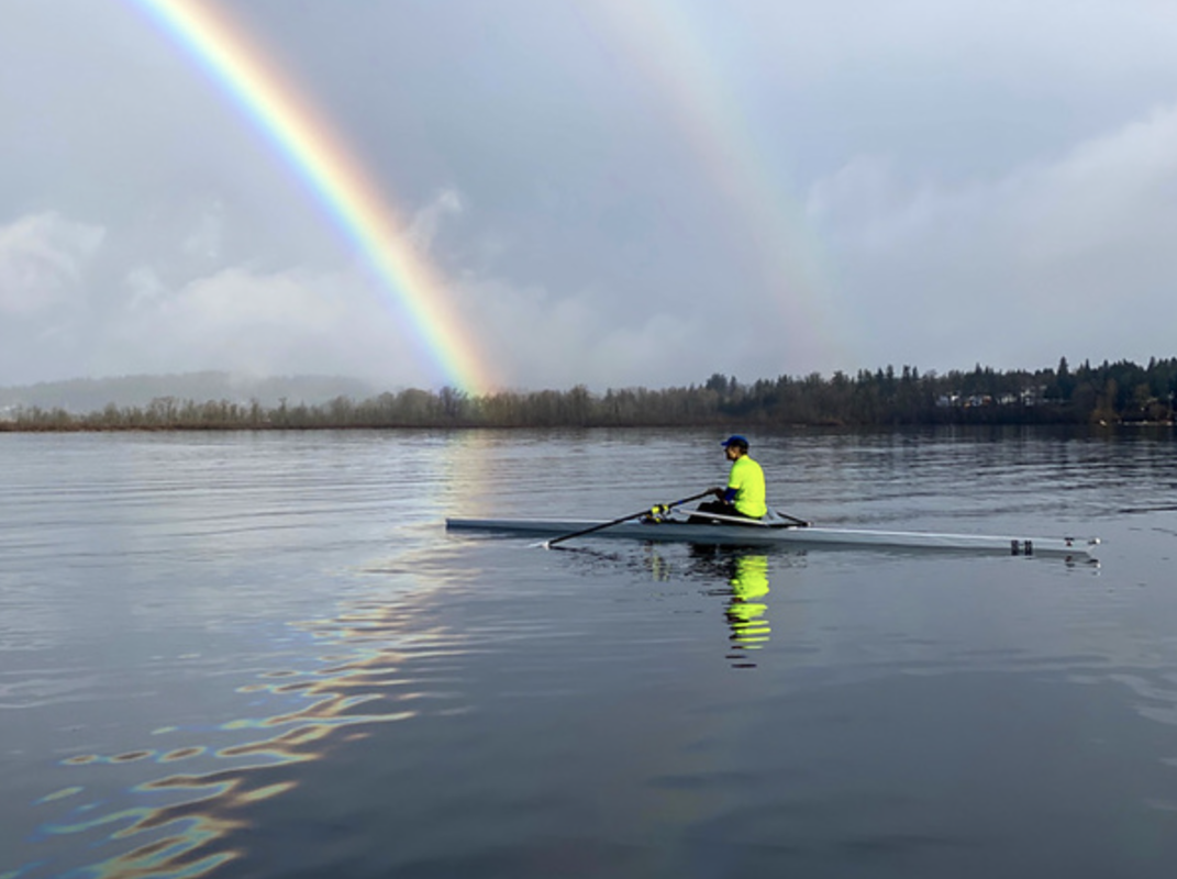 A rainbow is in the background of a photo of a single sculler wear bright neon yellow on Lake Sammamish
