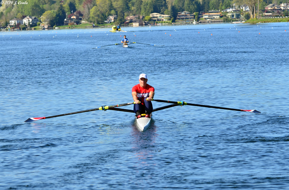 A rowing single is featured in the image foreground with two other singles in the background. They are rowing on a blue lake. The rower in the foreground wears a red SRA uniform with navy spandex pants. 
