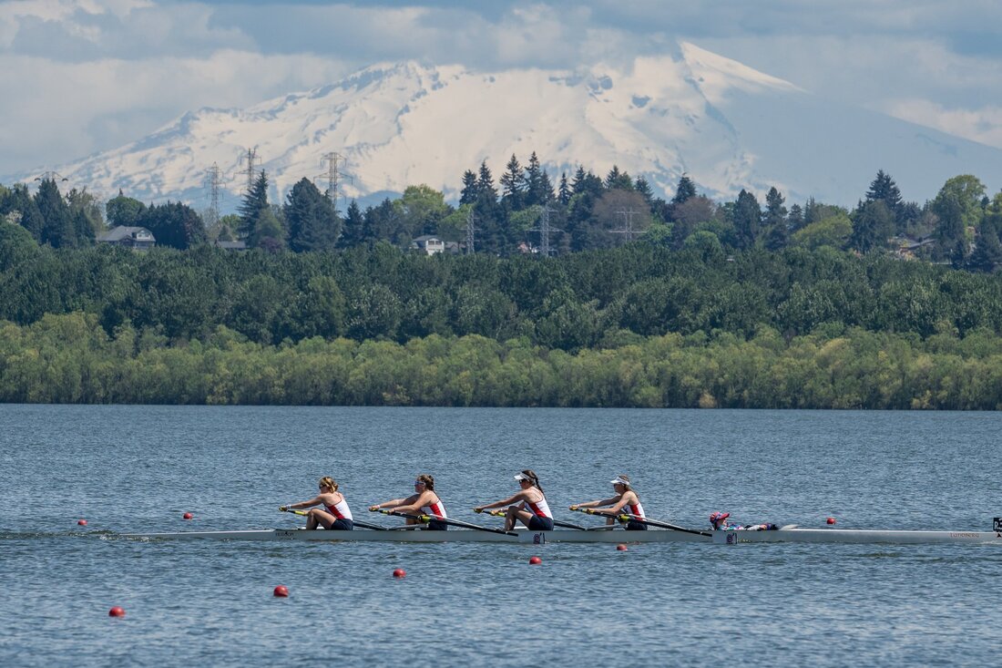 A junior Womens four rows by with a volcanic mountain in the background. They are rowing on Vancouver Lake and are wearing SRA unisuits. 