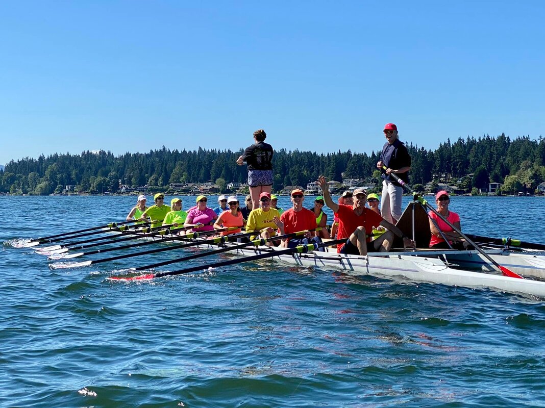 A rowing barge is full of masters rowers wearing neon yellow, pink, orange, yellow, and red shirts. They smile and wave at the camera as two coaches are standing in the center of the barge. The barge is floating on blue Lake Sammamish.