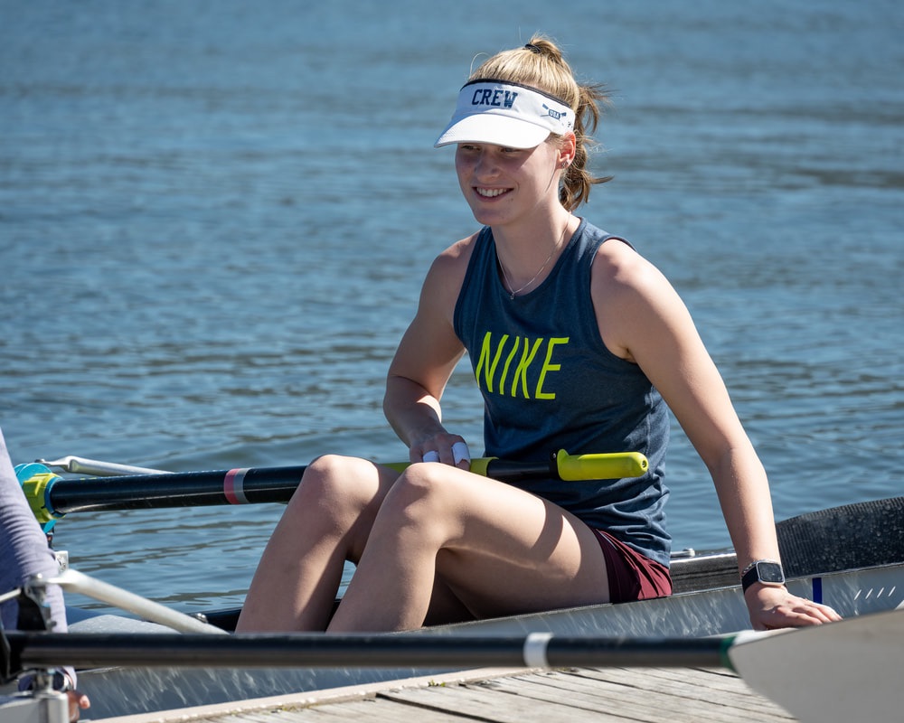 A rower from the junior girls team is smiling in the boat while grabbing onto the dock. She wear a tank top with the word 