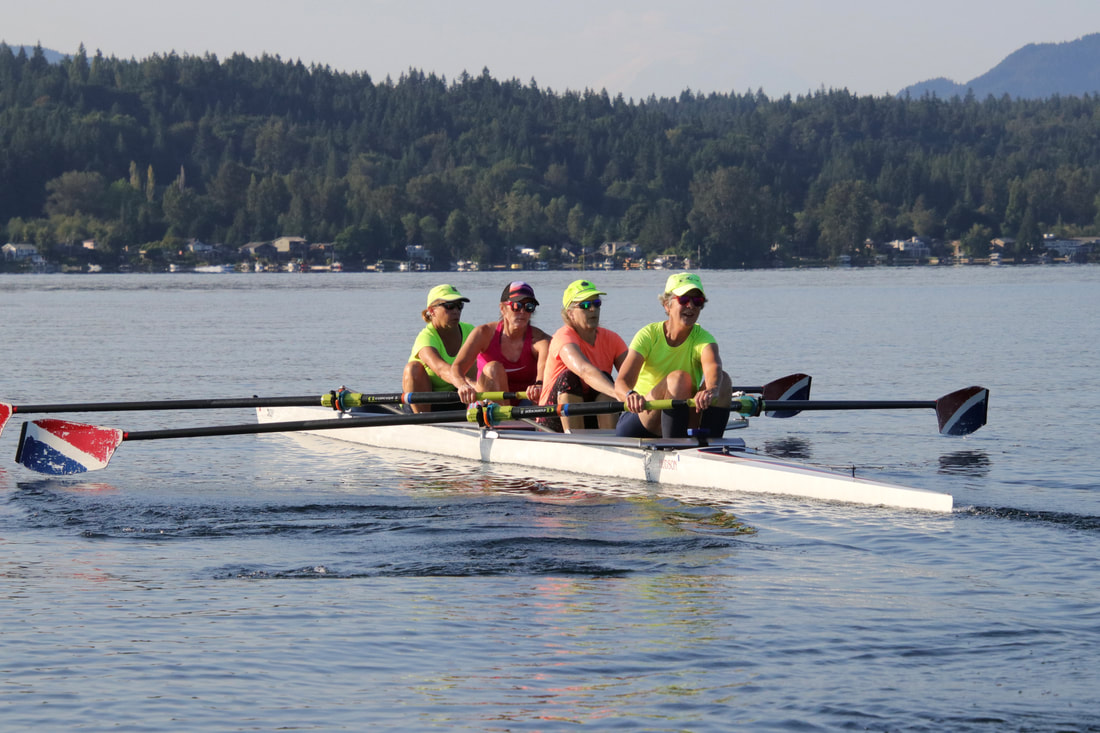 A Masters Four rows on Lake Sammamish with a mountain backdrop. The rowers wear bright neon clothes and their oars are navy, white, and red.