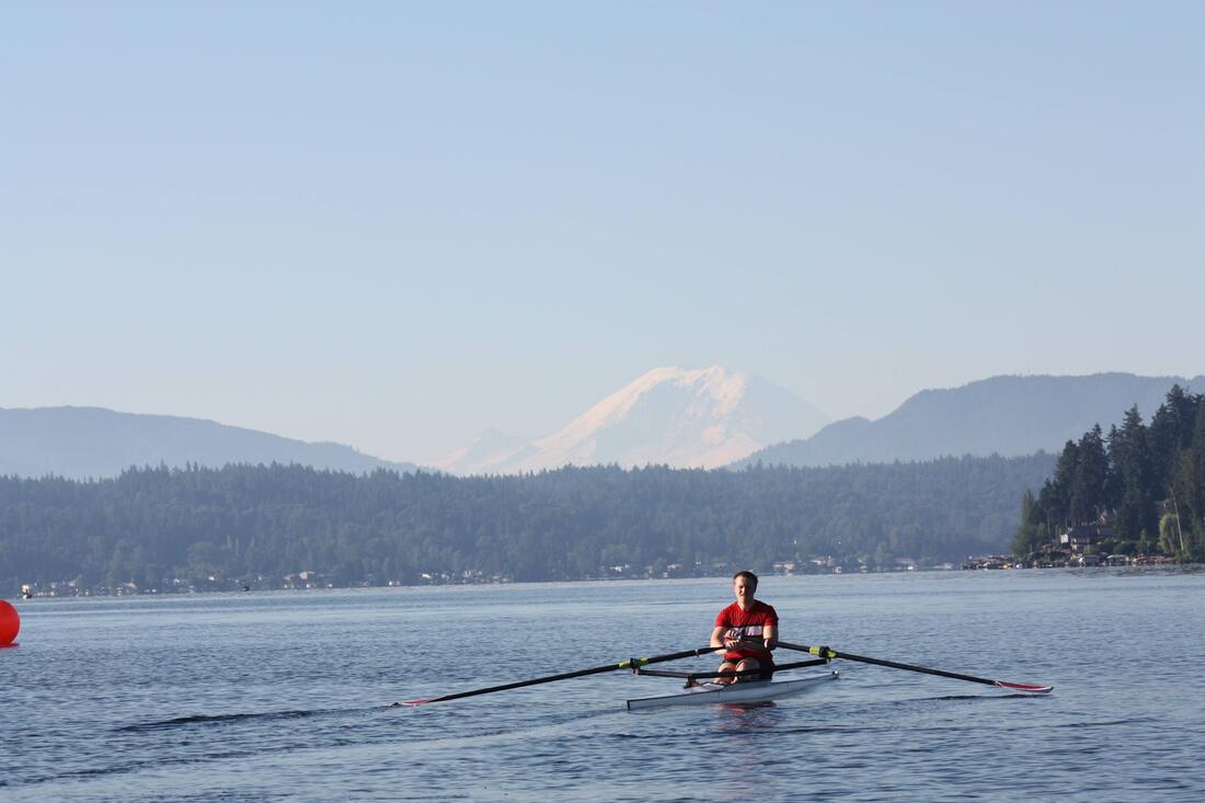A single with a man wearing a red shirt is seen on Lake Sammamish rowing towards Mount Rainier in the background. The sky is clear and the water is blue.