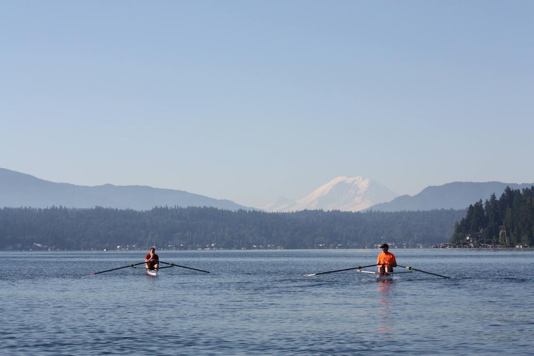 Two singles with a man wearing a red shirt and a man wearing an orange shirt row on Lake Sammamish heading towards Mount Rainier in the background