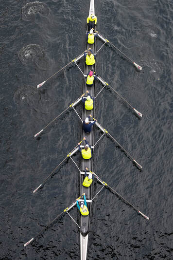 A rowing 8+ is pictured from an above shot and shows oarswomen in perfect unison. The water is dimpled by the puddles from the oars and the rowers wear neon yellow uniforms. 