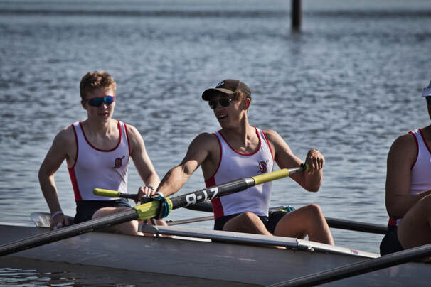 A rower leans back towards his teammate. The two are bow pair in what appears to be an eight. Both are holding their oars while keeping the boat set on water. 